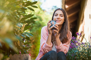 Relaxed young female smiling and looking at camera, sitting on home terrace, holding a mug drinking a hot beverage. Hobby and resting leisure concept.