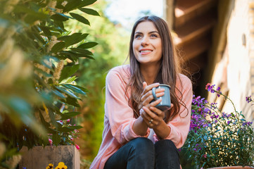 Happy young girl smiling and looking side, sitting on home terrace, holding a mug drinking a hot...