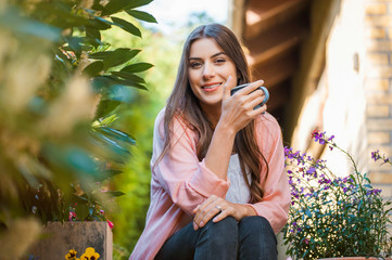 Pretty young girl smiling and looking at camera, sitting on home terrace, holding a mug drinking a hot beverage. Hobby and resting leisure concept.