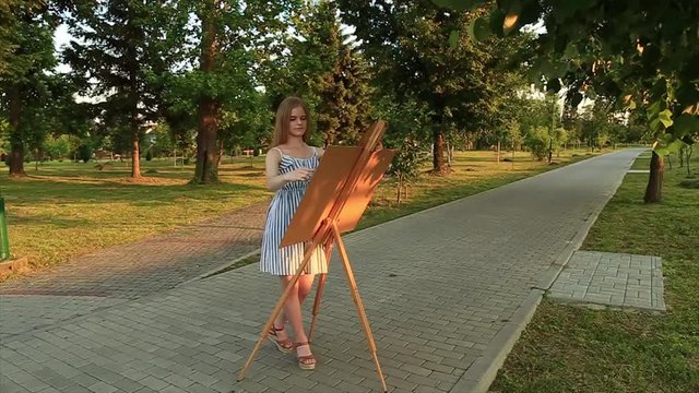 Beautiful girl standing in the park and draws a picture using a palette with paints and a spatula.
