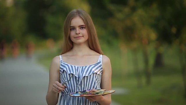 Beautiful girl artist is standing in the park and holding palette with paints