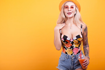 Fototapeta na wymiar Gorgeous blonde haired woman showing fresh orange juice with sunglasses and hat in studii over yellow background
