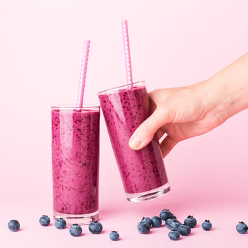 Two glasses of blueberries smoothie with straws on pink background. Woman's hand holding one glass. Healthy summer drink.