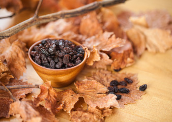 Black raisins in a wooden bowl. On dry leaf and brown wooden background.