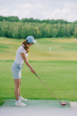 Fototapeta premium side view of woman in polo and cap playing golf at golf course