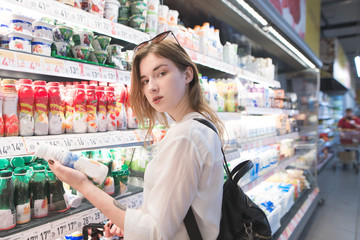 Young stylish woman standing in a yogurt supermarket in her hands and looking at the camera. Portrait of a woman who buys a yogurt in the milk department of a supermarket.