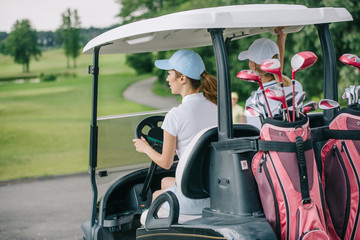 back view of female golfers in caps riding golf cart with equipment at golf course