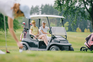 selective focus of woman playing golf while friends riding golf cart at golf course