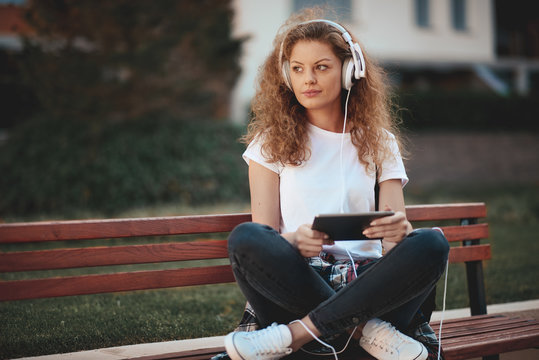 Young woman sitting on bench with legs crossed and listening to the music.