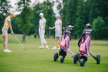 Fototapeta premium Selective focus of golf gear, woman in cap playing golf and friends standing near by at golf course