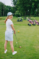 side view of smiling female golf player in cap with golf club standing at golf course