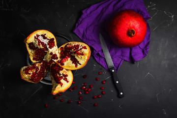 A whole ripe red pomegranate on a napkin. Pieces of a cut pomegranate, seeds, knife on a black background. Top view.