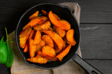 Organic food French fries, baked potatoes in rustic pieces in the skin. Ingredients for baked slices - ketchup, basil, sour cream sauce, salt, pepper. Dark black wooden background, pan.