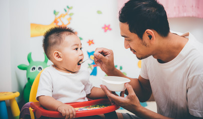 Obraz premium Father acting Mom feeding his son baby 1 year old on chair