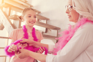 Like granny like granddaughter. Selective focus on a positive minded young lady smiling while standing next to her grandmother while both trying on bright pink feather boas.