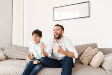 Photo of excited father 30s and son sitting together on sofa at home, and watching tv
