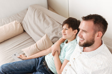 Portrait of joyful smiling father 30s and son sitting together on sofa at home, and looking aside