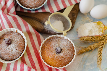 Cakes with cerries on a table in small porcelain cups .There are  eggs, flour, wheat, powdered sugar next to cakes.