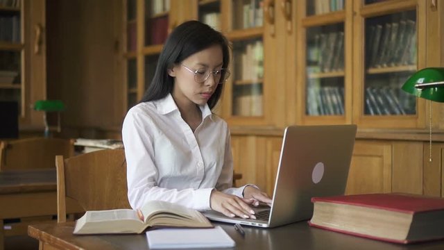 Beautiful woman is using laptop, reading book sitting at table in library, young brunette in white shirt is in educational process, researching subject indoors with bookcases. Concept: student