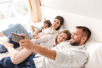 Photo of happy family with two kids smiling, and taking selfie on smartphone while lying together on bed in apartment