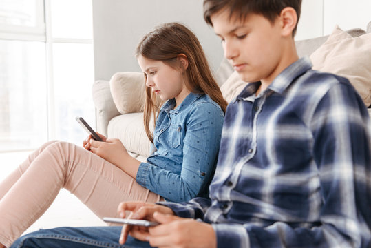 Image of two caucasian kids girl and boy sitting on floor near sofa at home, and both using mobile phone