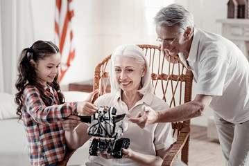 Look at it. Smiling grandma sitting in rocking chair carrying robot while happy girl and grandpa standing next to her and touching toy