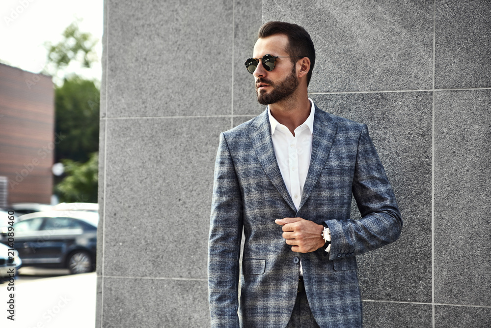 Wall mural Portrait of sexy handsome fashion businessman model dressed in elegant checkered suit posing near gray wall on street background. Metrosexual