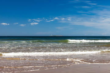 Lighthouse far out on the horizon in the middle with waves coming in on a beach