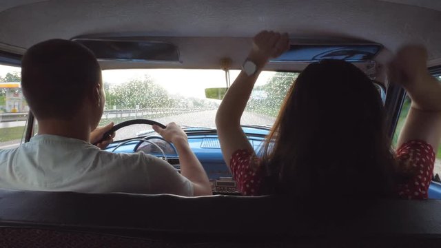 Young happy couple riding at vintage car and dancing. Unrecognizable man and woman sitting at the front seat of old retro car at road trip. Travel concept. Slow motion Rear back view