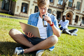 Watching video. Serious blonde man crossing legs while sitting on the grass and holding laptop on right hand