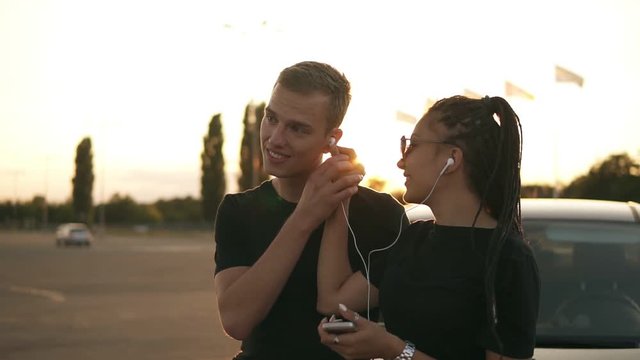 Two stylish friends or couple in black T shirts standing on a parking zone and having fun together while listening to the music using an earphones and smartphone. evening dusk