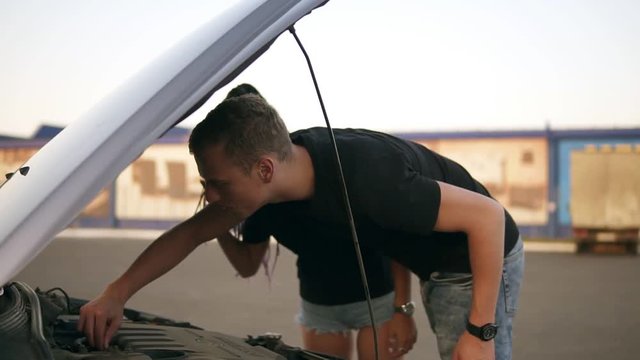 Young people on a roadtrip having a problem with their car, looking up for the failure with engine under the hood. Mall parking