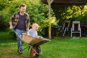 Happy little boy having fun in a wheelbarrow pushing by dad in domestic garden