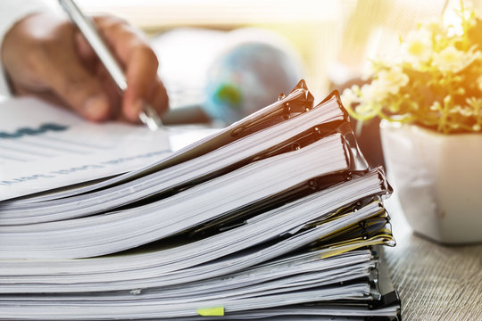 Businessman Hands Holding Pen For Working In Stacks Of Paper Files Searching Information Business Report Papers And Piles Of Unfinished Documents Achieves On Laptop Computer Desk In Modern Office