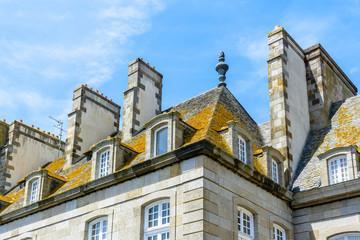 Fototapeta na wymiar View of the top of a residential building in the old city of Saint-Malo in Brittany, France, with a slate roof covered with lichen, dormer windows and large chimneys against blue sky.