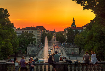 München bei Nacht - Blick auf die gut befahrene Prinzregentenstraße bei tollem Licht mit glücklichen Paaren im Vordergrund, die den goldenen Sonnenuntergang über der Stadt genießen - obrazy, fototapety, plakaty