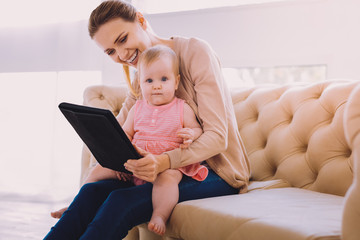 Progressive babysitter. Lovely baby girl sitting on the knees of a babysitter and looking at the pictures in her modern tablet