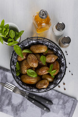 Baked potatoes in a black basket. Knife and fork. Olive oil, salt, pepper, basil. The background is white. Copy space. Top view. Vertical shot.