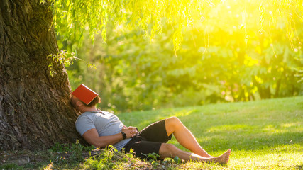 a young man is sleeping under the tree (weeping willow) with the book on his face