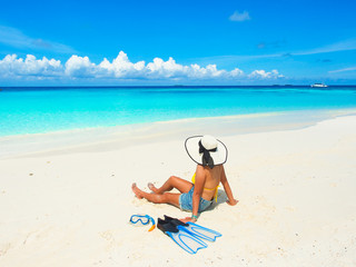 Fototapeta na wymiar Tanned woman wearing white hat sitting on the beautiful beach with snorkel and fins looking to turquoise sea in sunny day at Maldives island for travel or summer vacations holiday concept.