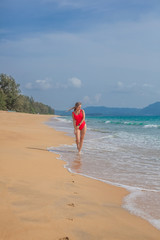 Sexy woman in red swimsuit strolls along the beach in water with small waves, Phuket, Thailand