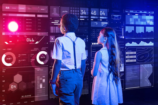 Two Modern Kids. Calm Clever Boy And Girl Standing In Front Of A Large Futuristic Device And Looking At The Transparent Screen Of It While Being Interested In The Construction Of A Security System