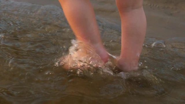 toddler's feet go on water on beach, joyful child stomps his feet in water, close-up, slow motion