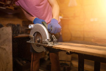 A young male carpenter-carpenter saws a modern circular saw with a wooden board in the workshop, wooden sawdust flying in the sides. to plan a tree.