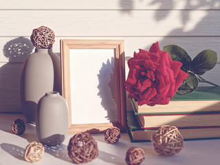 A red rose is on the books. Nearby there is an empty wooden frame. On a white table are the gray bottles and wicker balls. White wooden background.