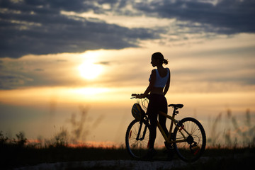 Fototapeta na wymiar Silhouette of sporty cyclist, wearing sportswear and posing near her bicycle on trail. Slender incognito woman enjoying nature and observing wonderful landscapes and amazing sunset.