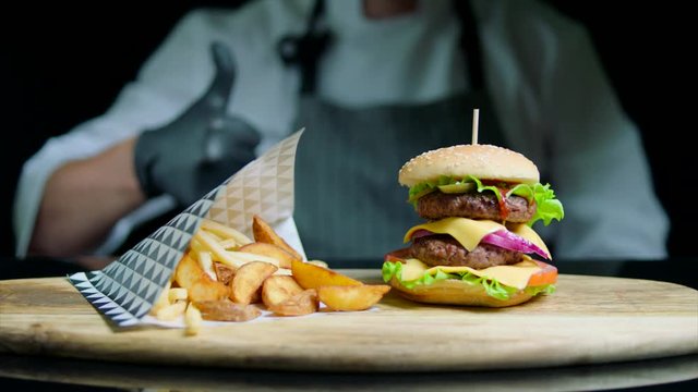 Chef is showing thumb out of focus and set of tasty burger, french fries, beer on black background
