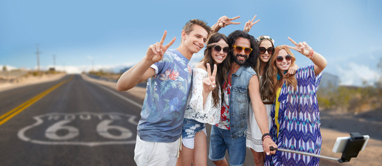 travel, technology and road trip concept - smiling young hippie friends taking picture by smartphone on selfie stick and showing peace hand sign over us route 66 background