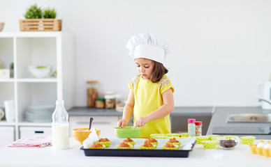 family, cooking, baking and people concept - little girl in chefs toque making batter for muffins or cupcakes at home kitchen