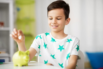 money, finances, childhood and people concept - smiling little boy putting coin into piggy bank at...